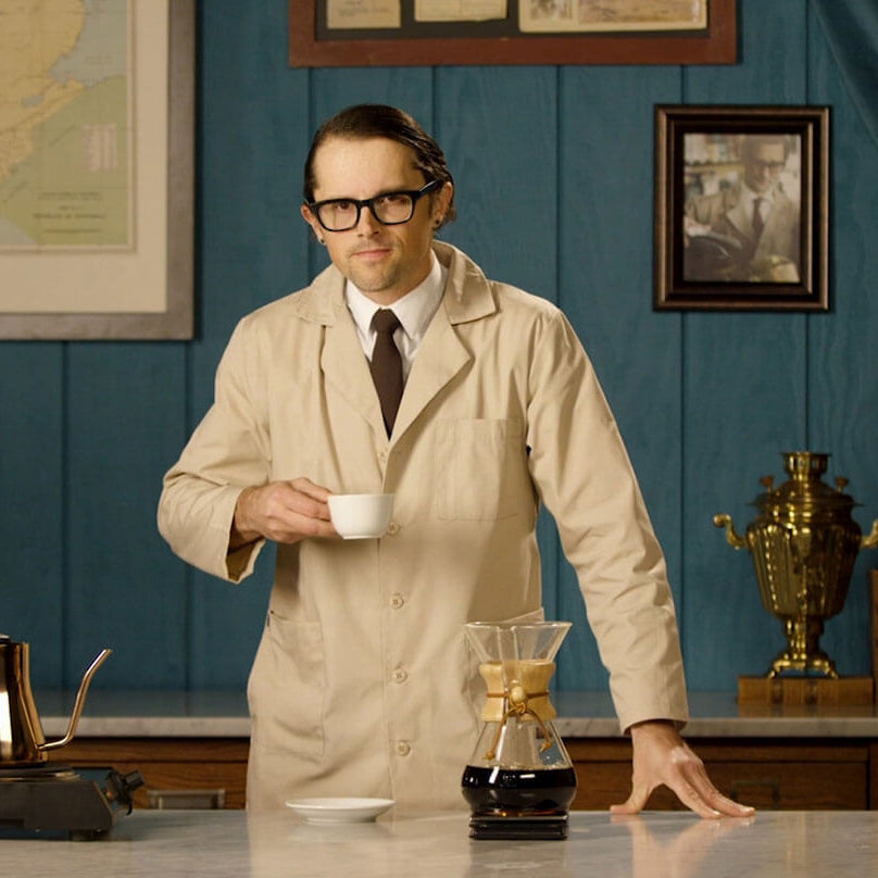 Man in a lab coat standing in a room with vintage decor, holding a cup.